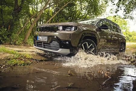 Das Foto zeigt einen Jeep Compass Elektro im Offroad-Einsatz bei einer Wasserdurchfahrt. Der kompakte Elektro-SUV fährt durch einen seichten Bach in bewaldeter Umgebung; die robuste Front mit markantem Jeep-Kühlergrill und erhöhter Bodenfreiheit ist deutlich zu sehen.