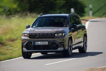 Das Foto zeigt einen Jeep Compass Elektro (Jeep Compass Electric) in Frontansicht auf einer kurvigen Landstraße. Der kompakte SUV mit Elektroantrieb fährt in leichter Kurvenlage; markant sind die rechteckigen LED-Scheinwerfer und der geschlossene Grillbereich. Die Szene vermittelt eine Straßenfahrt unter Tageslichtbedingungen in ländlicher Umgebung.