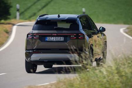 Das Foto zeigt den Jeep Compass Elektro aus der Heckansicht auf einer kurvigen Landstraße. Das elektrisch angetriebene Kompakt-SUV ist mit durchgehendem Rücklichtband, Dachspoiler und markanter Jeep-Schrift am Heck zu sehen und befindet sich in Kurvenfahrt. Die Szene spielt auf einer ländlichen Strecke mit Leitpfosten und Vegetation am Straßenrand.