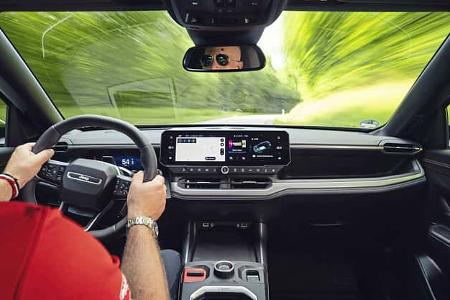 Der Innenraum des Jeep Compass Elektro ist aus Fahrerperspektive während einer Fahrszene zu sehen. Das Foto zeigt Multifunktionslenkrad mit Jeep-Logo, Digitalcockpit und zentrales Infotainmentsystem sowie die Mittelkonsole; durch die Frontscheibe ist die in Bewegung verschwimmende Landstraße erkennbar. Die IPTC-Angabe „Fahrszene“ bestätigt den fahrdynamischen Kontext.