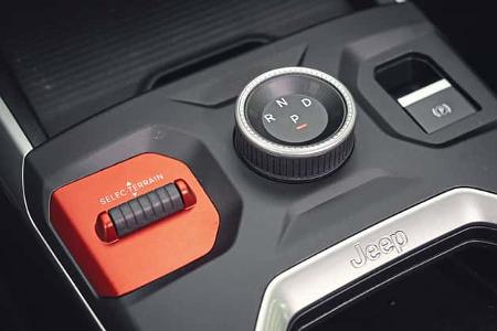 Die Detailaufnahme zeigt die Mittelkonsole des Jeep Compass Elektro. Sichtbar sind der drehbare Getriebewähler mit den Positionen P, R, N und D, der rote Selec‑Terrain‑Schalter für die Fahrmoduswahl sowie die Taste der elektrischen Parkbremse; der Jeep‑Schriftzug ist im Zierelement eingeprägt.
