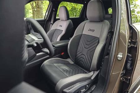 Das Foto zeigt den Innenraum eines Jeep Compass Elektro mit Fokus auf Fahrer- und Beifahrersitz. Die zweifarbigen Vordersitze tragen einen gestickten Jeep-Schriftzug und ein gemustertes Mittelteil; am Fahrersitz sind elektrische Verstellschalter sichtbar, daneben das Cockpit mit Multifunktionslenkrad und Mittelkonsole. Die Aufnahme erfolgt bei geöffneter Tür mit Blick ins grüne Umfeld durch die Scheiben.