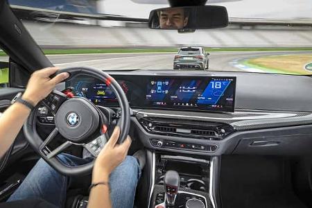 Das Foto zeigt den Innenraum eines BMW M2 Coupe aus der Fahrerperspektive während einer Fahrszene. Sichtbar sind das Cockpit mit gebogenem Curved-Display und digitalen Instrumenten, ein M-Lenkrad mit roten M-Tasten, Carbon-Zierleisten sowie ein Automatik-Wählhebel. Durch die Frontscheibe ist auf der Rennstrecke ein vorausfahrendes Fahrzeug zu sehen.
