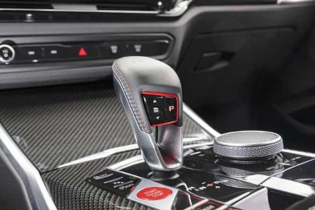Die Detailaufnahme zeigt die Mittelkonsole des BMW M2 Coupe mit dem Automatikwählhebel. Neben dem kurzen Wählhebel mit Park-Taste sind iDrive-Drehregler, Start‑Stopp‑Knopf und Bedientasten auf einer hochglänzenden, mit Carbonfaser-Zierleisten versehenen Oberfläche zu sehen.