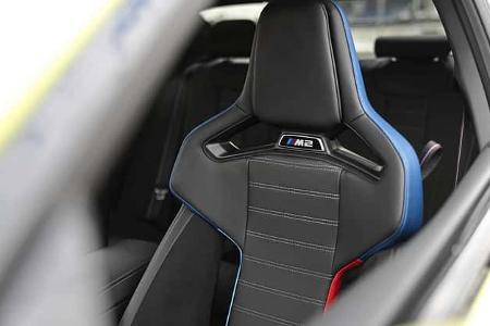 Das Foto zeigt den vorderen M-Sportsitz eines BMW M2 Coupe. Der Sportschalensitz verfügt über integrierte Kopfstütze, ausgeprägte Seitenwangen, einen Durchbruch in der Lehne sowie Kontrastnähte und blaue Zierpaspeln; in der Rückenlehne ist das M2‑Emblem sichtbar. Die Perspektive blickt durch die Seitenfensteröffnung in den Innenraum.
