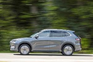 Das Foto zeigt den Geely E5 in vollständiger Seitenansicht während der Fahrt auf der Straße. Der graue SUV bewegt sich vor bewaldetem Hintergrund; durch den Mitzieher sind Räder und Umgebung verwischt, während die Karosserielinie und Fensterchromleisten klar erkennbar bleiben. Der Kontext entspricht einer Straßenfahrt ohne sichtbare Event- oder Ortsangabe.