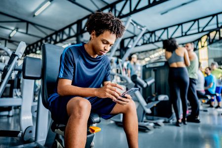Junger Mann sitzt im Fitnessstudio und schaut auf sein Handy