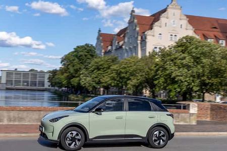 Das Foto zeigt einen Geely E2 Ultra in der Farbe Polarlichtgrün in klarer Seitenansicht. Der fünftürige Kompakt‑Crossover fährt auf einer innerstädtischen Straße; die geschlossene Front und aerodynamische Felgen deuten auf ein Elektroauto hin. Im Hintergrund sind Flussufer, Bäume und historische Stadtgebäude zu sehen.