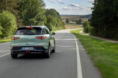 Das Foto zeigt einen Geely E2 Ultra in der Farbe Polarlichtgrün aus der Heckperspektive auf einer kurvigen Landstraße. Sichtbar sind die flachen Rückleuchten, das schwarze Dach und der GEELY-Schriftzug auf der Heckklappe. Die Aufnahme wirkt dynamisch und führt durch eine ländliche, von Wald und Feldern geprägte Umgebung.