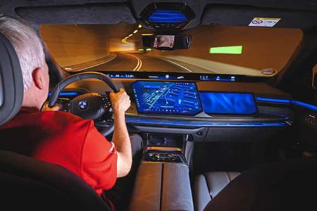 Das Foto zeigt den Innenraum eines BMW 740 xDrive während einer Fahrt durch einen Tunnel. Das Cockpit wird von einem breiten digitalen Displayverbund mit großem Navigationsdisplay und blau akzentuierter Ambientebeleuchtung dominiert; das BMW‑Lenkrad und Bedienelemente der Luxuslimousine sind deutlich erkennbar. Die Szene ist als Fahrszene im Tunnel aufgenommen.