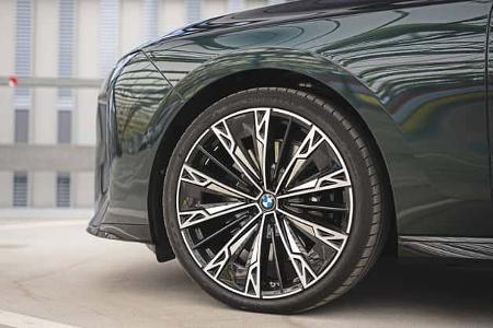 Die Detailaufnahme zeigt das Vorderrad der BMW 740 xDrive aus der 7er-Reihe. Sichtbar ist eine großformatige, mehrspeichige Leichtmetallfelge mit Niedrigprofilreifen und zentralem BMW‑Emblem; Reflexionen auf Kotflügel und Tür betonen das markante Felgendesign. Der Hintergrund wirkt urban und erinnert an ein Parkhaus mit strukturierter Fassade.