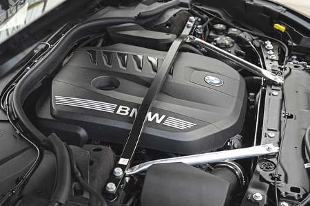 Die Aufnahme zeigt den Motorraum eines BMW 740 xDrive. Sichtbar sind die großflächige Motorabdeckung mit BMW-Schriftzug und Emblem sowie zwei Aluminium-Domstreben, dazu umliegende Leitungen und Bauteile der Vorderwagenstruktur. Die Detailansicht fokussiert den Antriebsbereich der Luxuslimousine.
