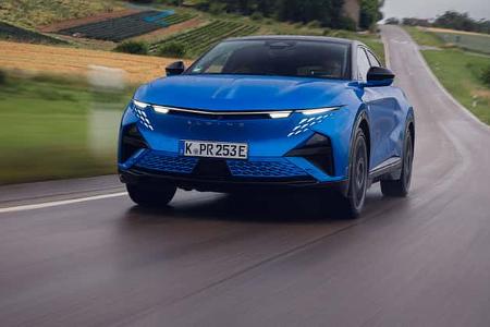Das Foto zeigt den Alpine A390 GT in Frontansicht auf einer nassen Landstraße. Der blaue Crossover/SUV fährt dynamisch; die schmale LED-Lichtleiste, die markante Tagfahrlicht-Signatur und die weitgehend geschlossene Frontpartie sind deutlich zu erkennen. Bewegungsunschärfe und Sprühwasser betonen die Fahrt auf der Straße.