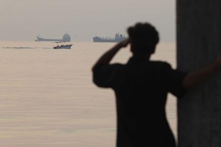 Ein Mann blickt aufs Wasser, während ein kleines Boot an Frachtschiffen vorbeifährt, die in der Straße von Hormus vor Bandar Abbas im Iran vor Anker liegen.