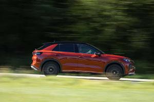 Das Bild zeigt einen VW T-Roc 1.5 eTSI in voller Seitenansicht während der Fahrt. Das kompakte SUV mit 1,5‑Liter eTSI‑Antrieb (Mildhybrid-Benziner) ist in einer dynamischen Mitzieher-Aufnahme festgehalten, die Bewegungsunschärfe im Hintergrund erzeugt. Die Szene spielt auf einer Straße vor grünem Bewuchs.