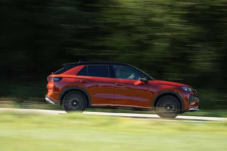 Das Bild zeigt einen VW T-Roc 1.5 eTSI in voller Seitenansicht während der Fahrt. Das kompakte SUV mit 1,5‑Liter eTSI‑Antrieb (Mildhybrid-Benziner) ist in einer dynamischen Mitzieher-Aufnahme festgehalten, die Bewegungsunschärfe im Hintergrund erzeugt. Die Szene spielt auf einer Straße vor grünem Bewuchs.