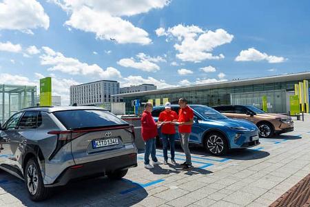 Die Aufnahme zeigt eine Gruppe von Elektro-SUVs im Vergleichstest – Toyota bZ4X, Opel Grandland und MG S6 EV – an markierten Ladeplätzen auf einem Parkdeck. Der Toyota bZ4X ist links mit angeschlossenem Ladekabel zu sehen, während drei Personen in roten Shirts neben den Fahrzeugen Unterlagen besprechen; die Szene wirkt wie eine Ladeszene im Testumfeld vor einem modernen Gebäudekomplex.