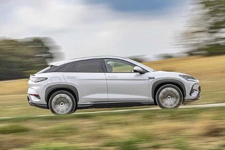 Das Foto zeigt einen BYD Sealion 7 EXcellence AWD in vollständiger Seitenansicht während der Fahrt. Das mittelgroße Crossover-SUV mit Allradantrieb ist in weißer Lackierung zu sehen; große Räder, schwarze Radlaufverkleidungen und eine fließende Dachlinie prägen das Exterieur. Die Aufnahme wurde als Mitzieher auf einer Landstraße in ländlicher Umgebung erstellt, was zu einem verwischten Hintergrund führt.