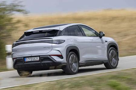 Das Foto zeigt den BYD Sealion 7 EXcellence AWD in Heck‑Dreiviertelansicht während der Fahrt auf einer Landstraße. Das coupéhaft gezeichnete Elektro‑SUV mit Allradantrieb trägt ein deutsches E‑Kennzeichen; das durchgehende Rücklichtband und der schwarze Dachspoiler sind deutlich erkennbar. Der Wagen ist in silberner Lackierung in ländlicher Umgebung unterwegs.