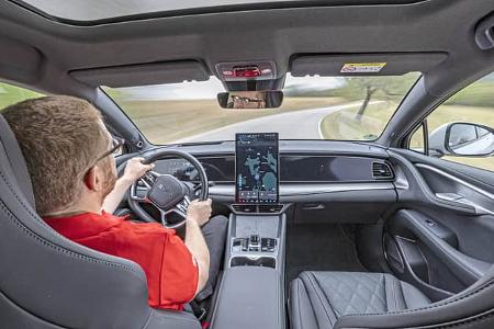 Die Aufnahme zeigt den Innenraum des BYD Sealion 7 EXcellence AWD aus der Perspektive der Rücksitzbank während der Fahrt. Sichtbar sind das BYD‑Lenkrad mit digitalem Kombiinstrument, ein großes hochformatiges Zentraldisplay mit Navigationsansicht, die breite Armaturentafel und die Mittelkonsole; Lederpolsterung und ein Panoramadach sind erkennbar. Die Szene spielt auf einer kurvigen Landstraße, der Fahrer hält das Fahrzeug am Steuer.