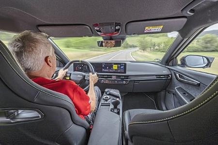 Die Aufnahme zeigt den Innenraum des Kia EV6 GT AWD aus der Rücksitzperspektive während der Fahrt auf einer Landstraße. Zu sehen sind das breit angeordnete Digitalcockpit mit Infotainmentsystem, ein Sportlenkrad sowie die Mittelkonsole mit Drehregler für die Fahrstufen; sportliche Sitze mit Kontrastnähten rahmen die Szene. Die GT-Variante mit Allradantrieb wird hier aus Fahrerperspektive dokumentiert.