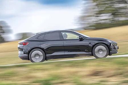 Das Bild zeigt einen Polestar 4 Dual Motor in Seitenansicht während der Fahrt auf einer Landstraße. Das vollelektrische SUV-Coupé mit zwei Elektromotoren (Allradantrieb) ist in Schwarz zu sehen; die fließende Dachlinie, bündige Türgriffe und aerodynamische Räder sind deutlich erkennbar. Die Bewegungsunschärfe im Hintergrund betont die Straßenfahrt in ländlicher Umgebung.