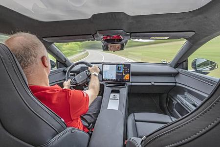 Das Foto zeigt den Innenraum des Polestar 4 Dual Motor aus der Rückbankperspektive während der Fahrt. Sichtbar sind das Polestar-Lenkrad, ein großes zentrales Touchscreen-Infotainmentsystem mit Navigationsanzeige, die breite Mittelkonsole sowie sportliche Vordersitze. Ein durchgehendes Glasdach und die klare, horizontale Cockpitgestaltung prägen den Eindruck.