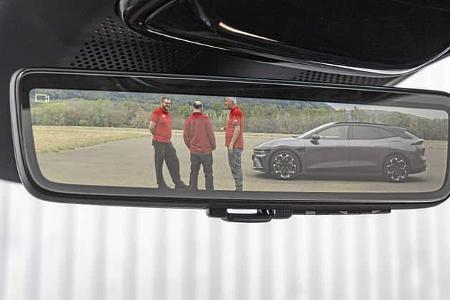 Das Foto zeigt den digitalen Innenspiegel eines Polestar 4 Dual Motor. Der spiegelintegrierte Monitor blendet das Kamerabild ein, in dem drei Personen sowie die seitliche Ansicht eines Polestar 4 auf einer asphaltierten Fläche zu sehen sind. Die Aufnahme fokussiert das Innenraum‑Detail und die Funktion des kamerabasierten Rückspiegels.