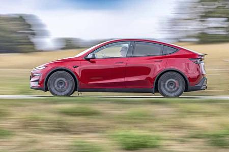 Das Foto zeigt ein Tesla Model Y Performance in voller Seitenansicht während der Fahrt. Die Mitzieher-Aufnahme betont die Bewegung; das rote Elektro-SUV mit coupéhafter Dachlinie und großen Rädern fährt auf einer Landstraße in ländlicher Umgebung, rote Bremssättel sind sichtbar.
