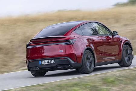 Das Foto zeigt einen Tesla Model Y Performance in roter Lackierung aus der Heck-Dreiviertelansicht auf einer kurvigen Landstraße. Als Performance-Variante des Elektro-SUV ist das Fahrzeug in Fahrt zu sehen, mit dunklen Leichtmetallrädern und getöntem Glasdach; die coupéartige Dachlinie wird deutlich hervorgehoben.