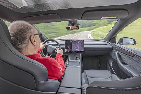 Der Innenraum eines Tesla Model Y Performance ist aus der Fahrerperspektive zu sehen. Das minimalistische Cockpit mit großem zentralen Touchscreen zeigt die Navigation, dazu sind Panoramadach, Sportsitze und die durchgehende Zierlinie am Armaturenbrett erkennbar. Die Aufnahme entsteht während der Fahrt auf einer kurvigen Landstraße.