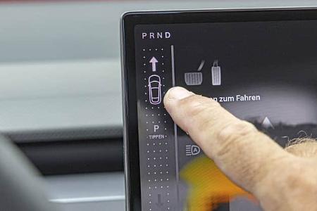 Die Detailaufnahme zeigt das zentrale Touchscreen-Display des Tesla Model Y Performance im Innenraum. Über die digitale Benutzeroberfläche erfolgt die Gangwahl (PRND) per Tippen, eine Hand interagiert gerade mit dem Menü; Symbole und Hinweise sind auf dem Infotainment-Display sichtbar. Die Szene fokussiert die HMI-Bedienlogik des Elektro-SUVs im Cockpit.