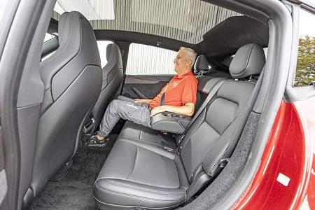Das Foto zeigt den Fond eines Tesla Model Y Performance; eine sitzende Person demonstriert den Platz auf der Rücksitzbank bei geöffneter Tür. Sichtbar sind schwarze Sitze mit klappbarer Mittelarmlehne, ein nahezu ebener Fahrzeugboden und das durchgehende Panoramaglasdach.
