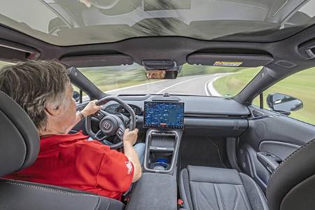 Der Innenraum des Zeekr 7GT AWD ist aus der Rücksitzperspektive während der Fahrt zu sehen. Das Cockpit zeigt ein großes zentrales Infotainment-Display mit Navigationskarte, ein digitales Kombiinstrument, eine breite Mittelkonsole und ein durchgehendes Panoramaglasdach; das gezeigte Modell ist die Allradantriebsvariante (AWD). Außen ist eine kurvige Landstraße erkennbar.