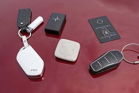 Das Foto zeigt verschiedene Fahrzeugschlüssel und Keyfobs der in den IPTC-Daten genannten Modelle Kia EV 6 GT AWD, BYD Sealion 7 EXcellence AWD, Polestar 4 Dual Motor, Tesla Model Y Performance und Zeekr 7GT AWD. Abgebildet sind klassische Funkschlüssel, kompakte Transponder und eine NFC‑Schlüsselkarte, arrangiert auf einer roten Karosserieoberfläche; die Designs unterstützen Keyless‑Go und kontaktlosen Fahrzeugzugang.