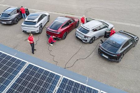 Fünf Elektro-SUVs – Kia EV6 GT AWD, BYD Sealion 7 Excellence AWD, Polestar 4 Dual Motor, Tesla Model Y Performance und Zeekr 7GT AWD – stehen nebeneinander und sind über Ladekabel angeschlossen. Im Vordergrund sind Photovoltaik-Module zu sehen; mehrere Personen prüfen den Ladevorgang auf einer großen asphaltierten Fläche.